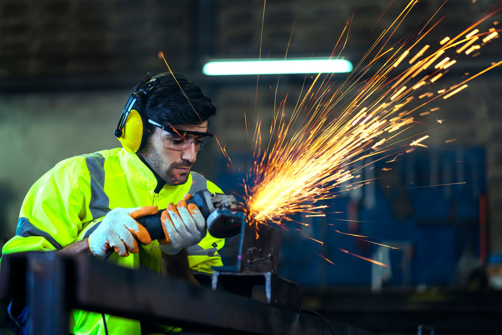 Trabajador masculino caucásico en un uniforme de pieles soldadores de desgaste safty, amolado de rueda eléctrica en la estructura de acero en factor en el garaje, chispas bokeh naranja vuelan a los lados